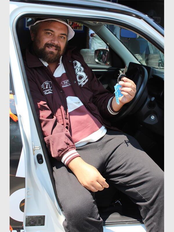 A man wearing a white bucket hat and brown jacket with a beard and moustache sitting in the driver's seat of a car holding the key.