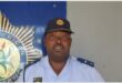 A black police officer in uniform, wearing a cap and standing against a wall with the SAPS badge.