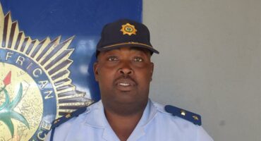 A black police officer in uniform, wearing a cap and standing against a wall with the SAPS badge.