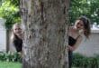 Two teenage girls wearing black looking out from behind a tree