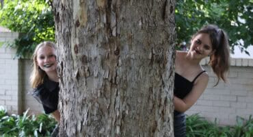 Two teenage girls wearing black looking out from behind a tree