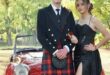 Boy wearing a kilt with a girl wearing a black dress pose in front of a red vintage car