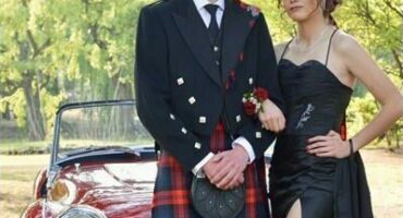 Boy wearing a kilt with a girl wearing a black dress pose in front of a red vintage car