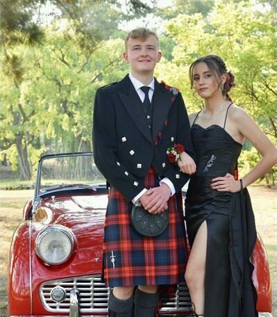 Boy wearing a kilt with a girl wearing a black dress pose in front of a red vintage car
