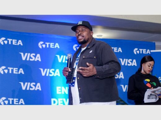 Black male with a microphone in his hand wearing a black cap and with a VISA banner in the background.