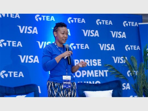 Black female wearing a blue top and holding a microphone with a VISA banner in the background. 