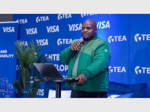 Black male wearing a green jacket and holding a microphone with a Visa backdrop in the background.