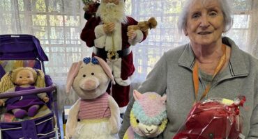Elderly woman with grey hair standing with plush toys and a Santa Claus statue in the background.