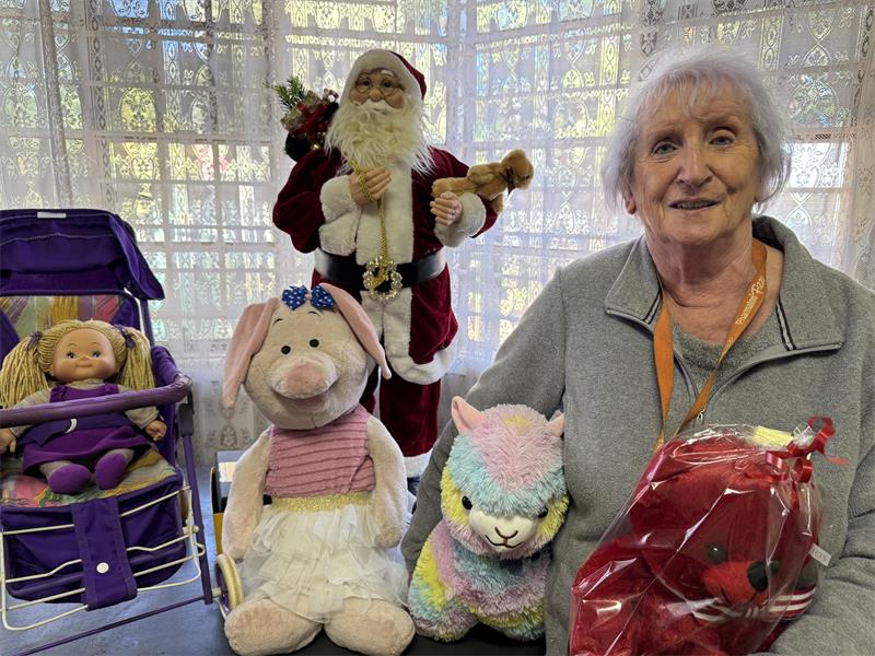 Elderly woman with grey hair standing with plush toys and a Santa Claus statue in the background.
