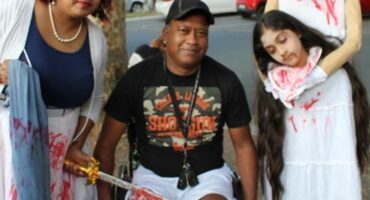 A photo of a woman covered in fake blood, holding a sword to a wheelchair-bound man’s leg with a headless girl standing beside him.