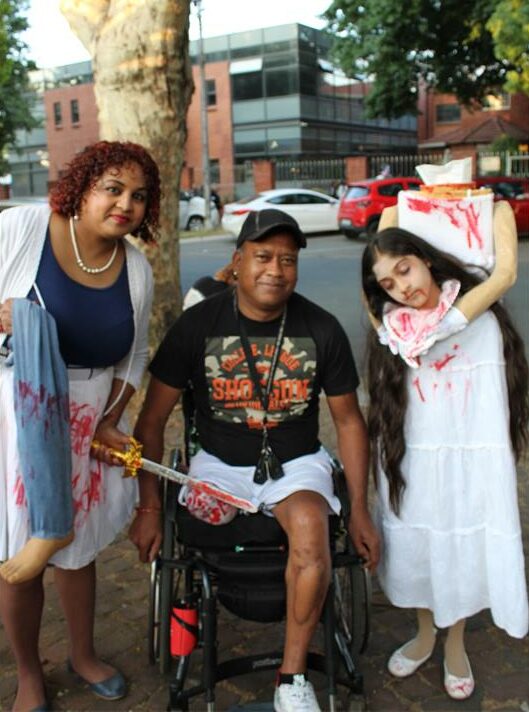 A photo of a woman covered in fake blood, holding a sword to a wheelchair-bound man’s leg with a headless girl standing beside him.