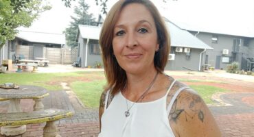 A woman with tattoos and long hair, posing in a garden setting.
