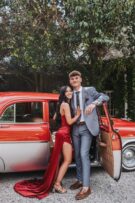 Girl wearing a red dress with a boy wearing a grey suit posing in front of a vintage red car
