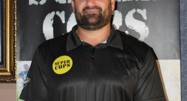 Man with a beard and black shirt with a yellow circle on the chest and Super Cops written in black.