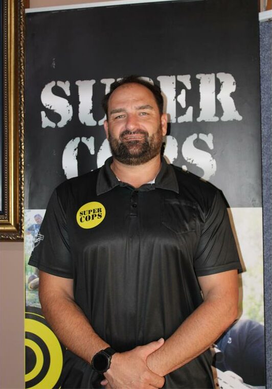 Man with a beard and black shirt with a yellow circle on the chest and Super Cops written in black.