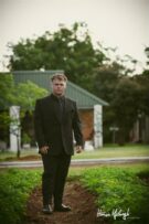 Young man in a black suit posing in a garden setting.