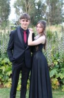 Boy wearing a black suit with a red tie standing next to a girl with a black dress with flowers in the background
