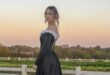 Girl in a black dress with white detailing standing in front white fence at sunset