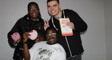 Woman in black stands with a young man holding a book with a white cover and a wheelchair-bound man holding a crochet chicken.