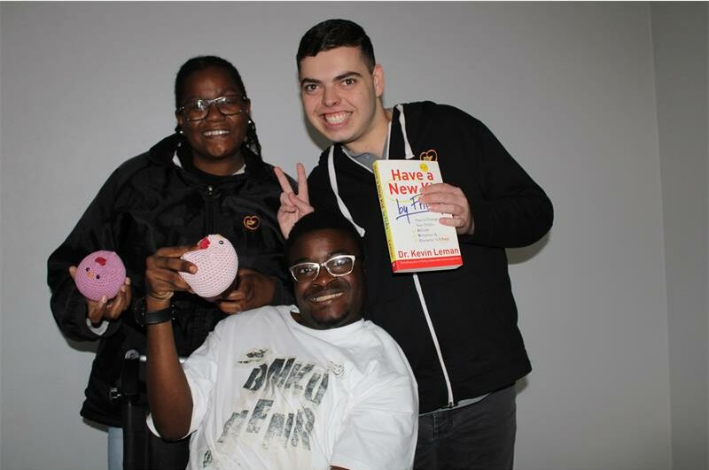 Woman in black stands with a young man holding a book with a white cover and a wheelchair-bound man holding a crochet chicken.