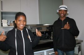 Girl in black hoodie and man with white bucket hat, standing in front of a coffee machine, showing a thumbs up.