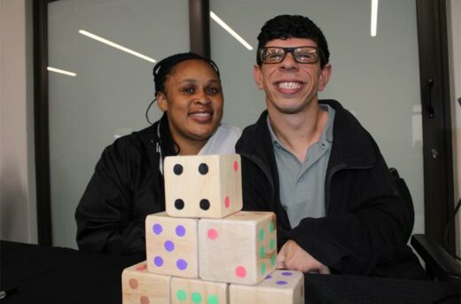 Black woman sitting next to a man with glasses. In front of them are six wooden dice.