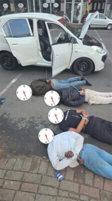 Four men lying in the road with a white vehicle in the background