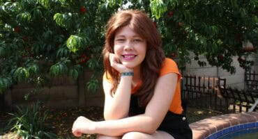 Girl wearing an orange top and black dress sits in front of a tree wearing a brown wig