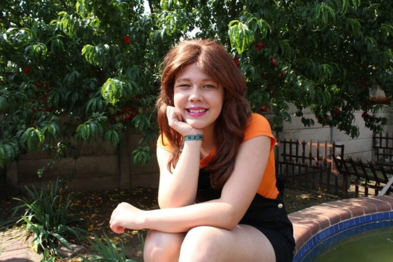 Girl wearing an orange top and black dress sits in front of a tree wearing a brown wig