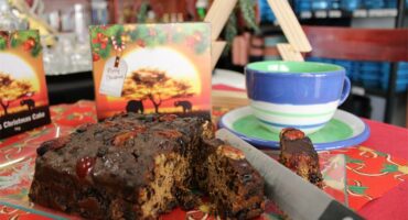 Bread knife cutting into a fruit cake with a cup and packaging in the background