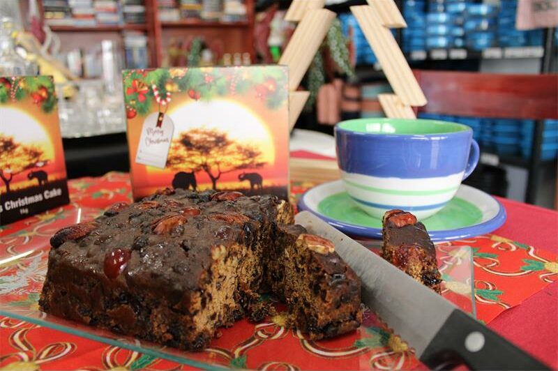 Bread knife cutting into a fruit cake with a cup and packaging in the background