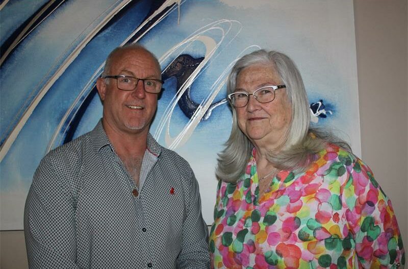 Man with glasses and woman in floral shirt standing in front of a blue background.