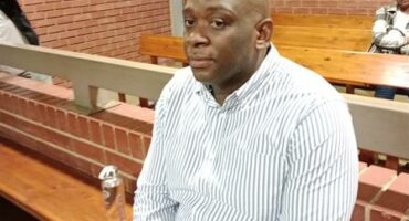 Man sitting in the dock in a court setting with his fingers crossed between his legs