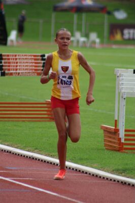 A photo of a girl wearing a yellow, white and red kit. She's running next hurdles on a red tartan tack.