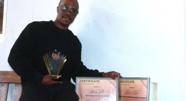 A photo of a man dressed in black. He's carrying a trophy and two certificates.