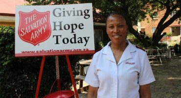 Woman wearing a white overall, standing next to a red and white Salvation Army sign.