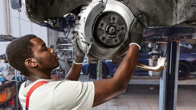 A black man working on a brake drum under a car.
