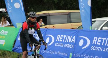 A photo of cyclist riding next a blue banner with the words Retina South Africa.