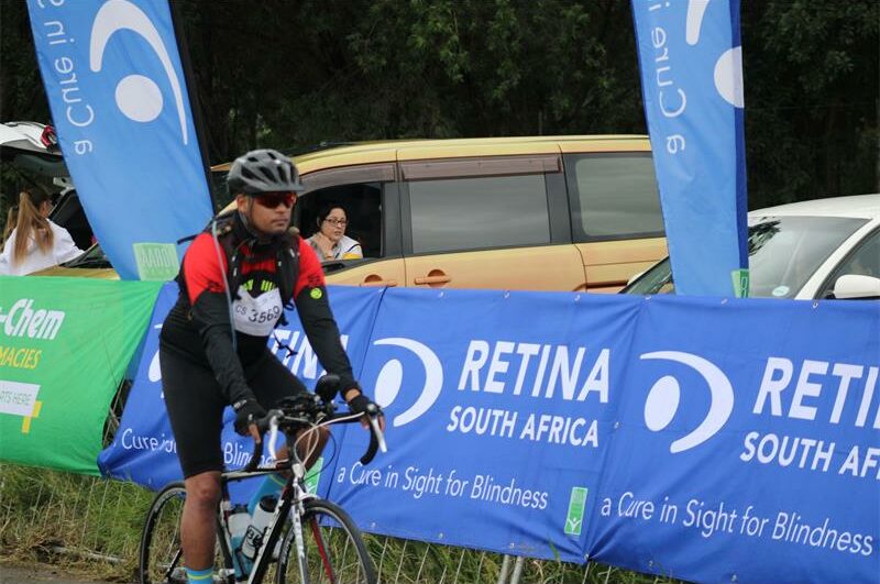 A photo of cyclist riding next a blue banner with the words Retina South Africa.