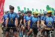 A photo of a group of cyclists wearing blue vests and helmets. They are on their bikes and there are green banners behind them.