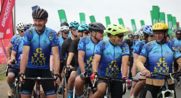 A photo of a group of cyclists wearing blue vests and helmets. They are on their bikes and there are green banners behind them.