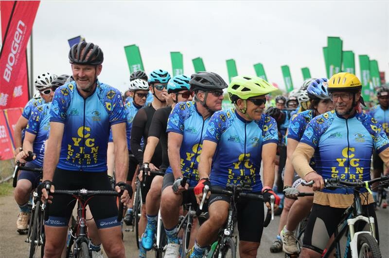 A photo of a group of cyclists wearing blue vests and helmets. They are on their bikes and there are green banners behind them.