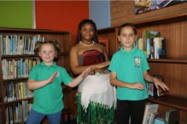 Two girls wearing green golf shirts dance with lady dressed in a Polynesian outfit