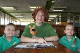 Boy holding a fluffy toy dog sits in front of a book with a boy and girl wearing green shirts. Photo: Jani de Beer