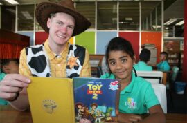 Indian girl wearing a green polo shirt sits with a cartoon character holding a book