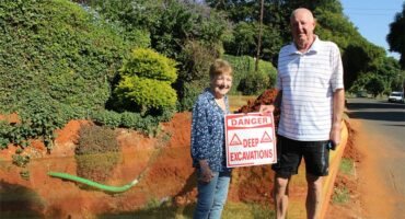 Elderly woman and man standing next to a hole holding a red and white sign