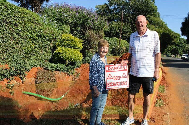Elderly woman and man standing next to a hole holding a red and white sign
