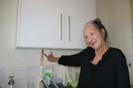 An elderly woman points to rust on a wall.
