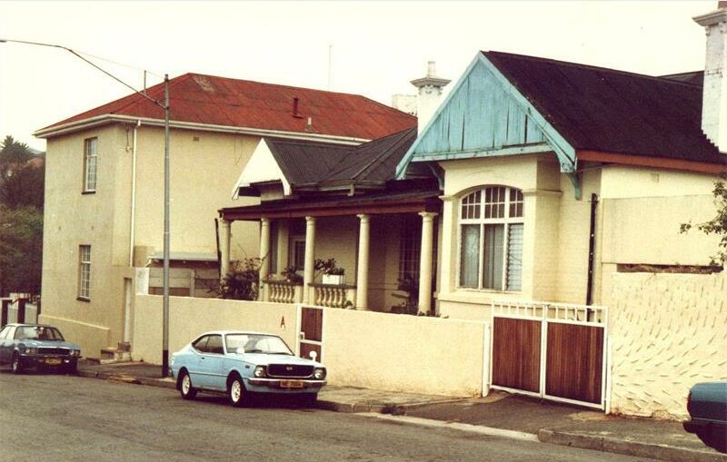 A colour photo of an old house and a blue old car parked on the street in front of it.