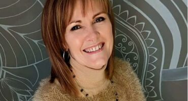 A white woman with ginger hair wearing a camel coloured sweater and necklace smiling at the camera with a grey and white patterned background.